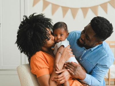 A Young Black couple smiling while kissing and holding their baby.