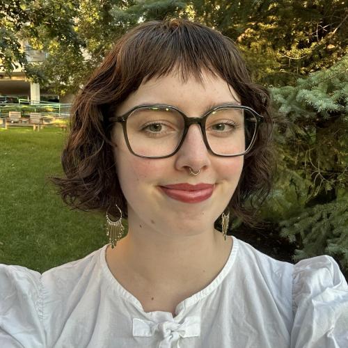 Nadia, wearing white and glasses - smiling in front of a tree